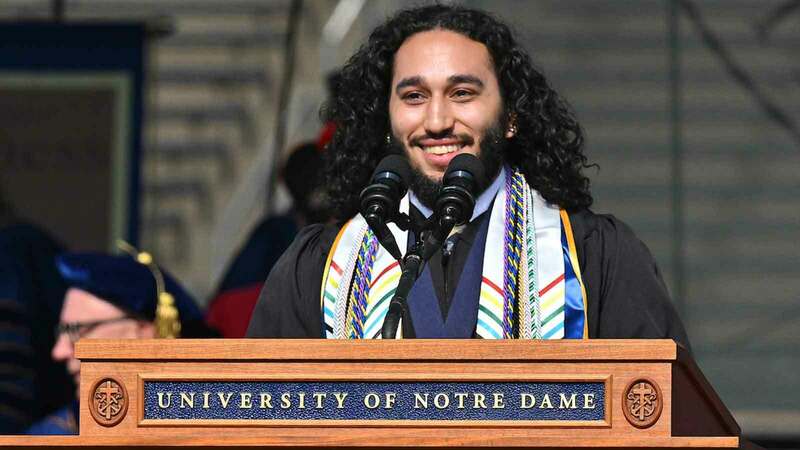 A young man with long black hair standing behind a University of Notre Dame podium at commencement.